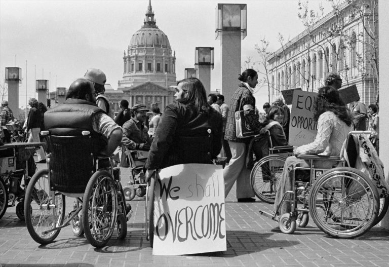 An old photo of people using wheelchairs who are advocating for their rights. A sign is attached to the back of a wheelchair that reads "We Shall Overcome".