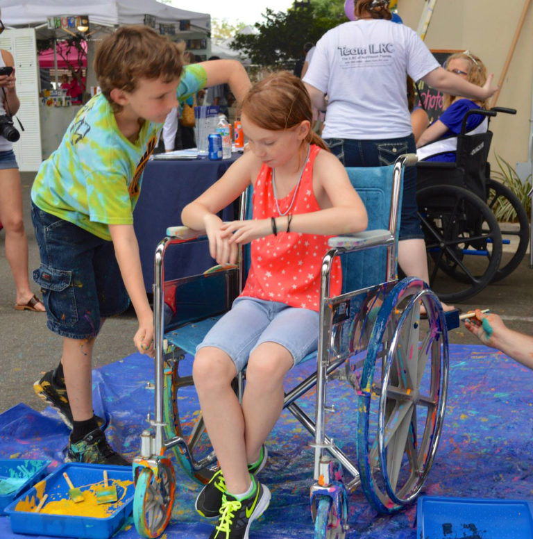 Two children, one sitting in a wheelchair, get ready to roll over canvas with paint