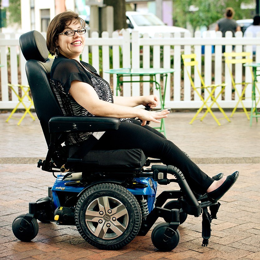 A smiling woman with glasses sits in a power wheelchair on a brick plaza.