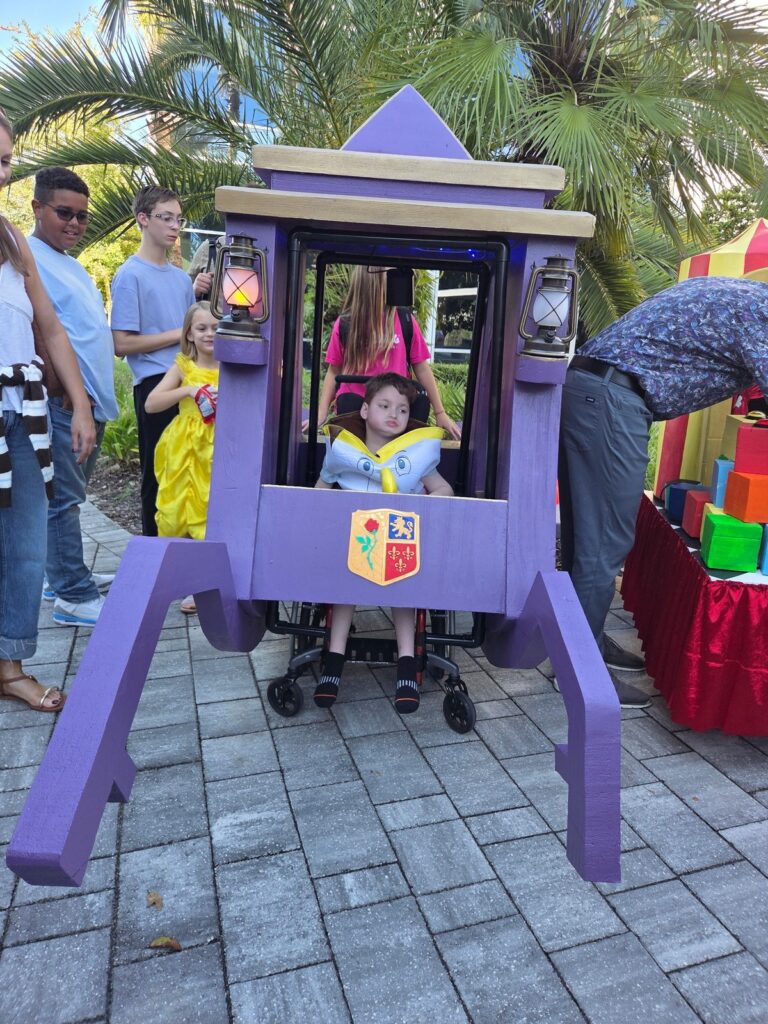 A boy in a wheelchair sits inside a purple carriage costume with lanterns, while children and adults stand nearby