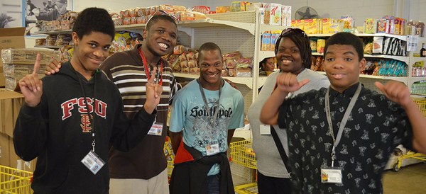 Five smiling young people in a food pantry wave and make hand signs at the camera.