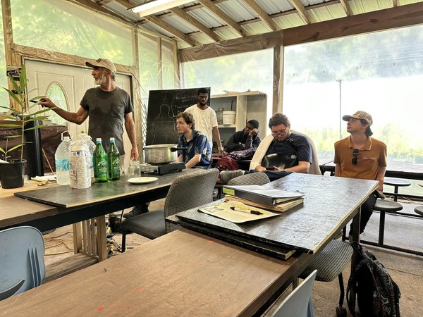A man speaks to a group seated around tables in an open-sided workshop.