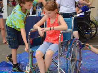 Two children, one sitting in a wheelchair, get ready to roll over canvas with paint