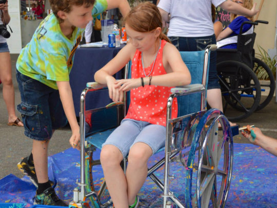 Two children, one sitting in a wheelchair, get ready to roll over canvas with paint