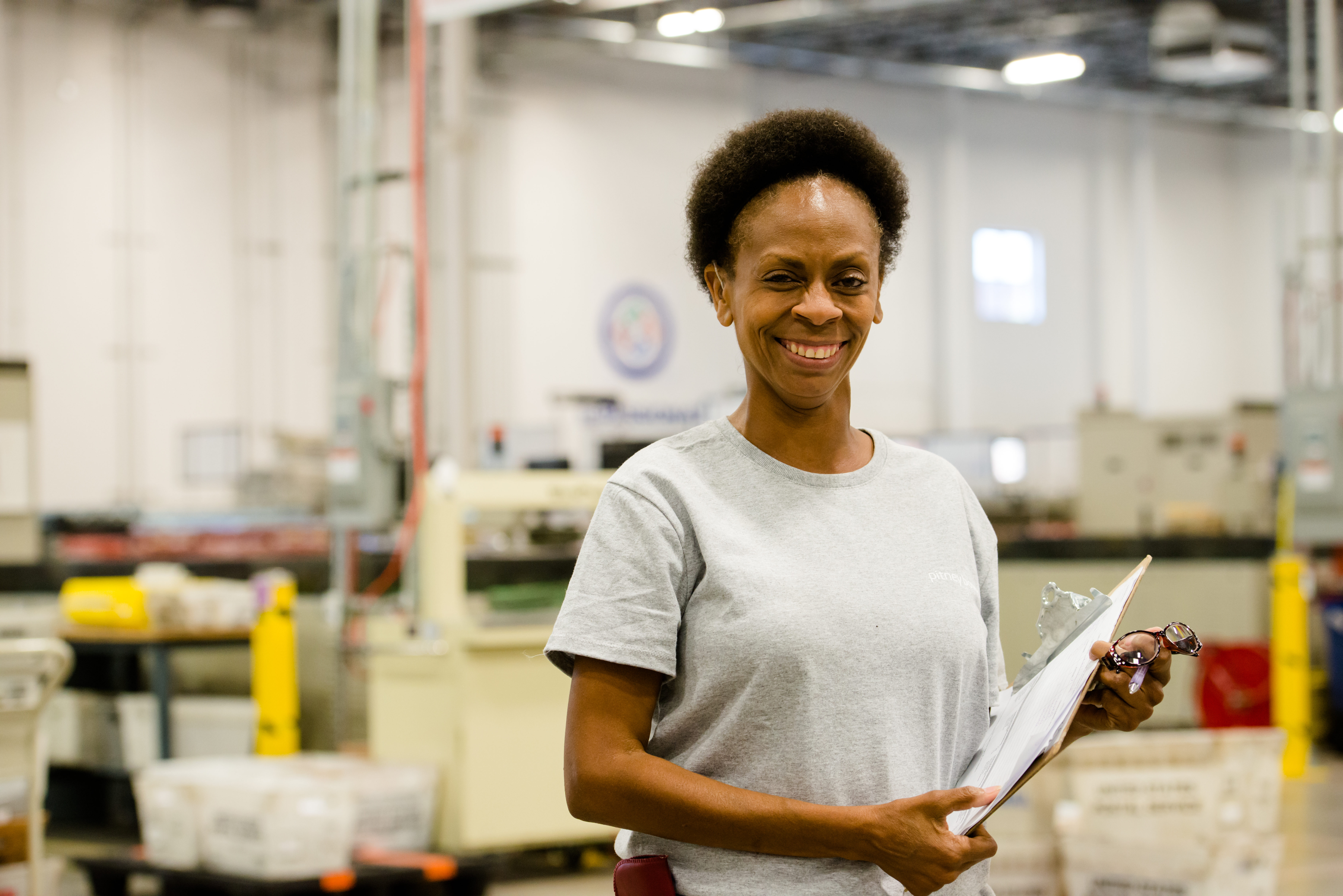 A woman holding a clipboard and smiling. A factory is in the background.