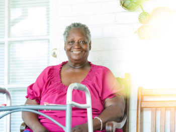 A woman in a pink shirt smiles. A walker is in the foreground.