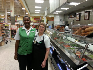 Two women who work at Publix, in uniform, one in a regular vest and the other in an apron. They have an arm around each other next to a deli inside Publix.