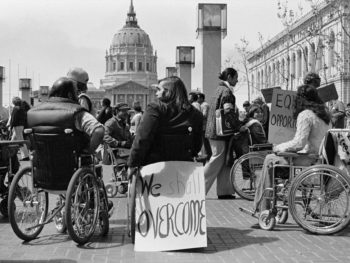 An old photo of people using wheelchairs who are advocating for their rights. A sign is attached to the back of a wheelchair that reads 